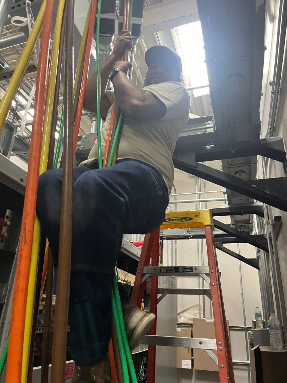Technician climbing a ladder while managing colorful electrical cables in a workspace.