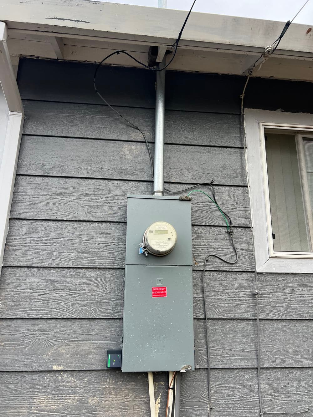 Electric meter mounted on a gray exterior wall, with wires and a warning label visible.