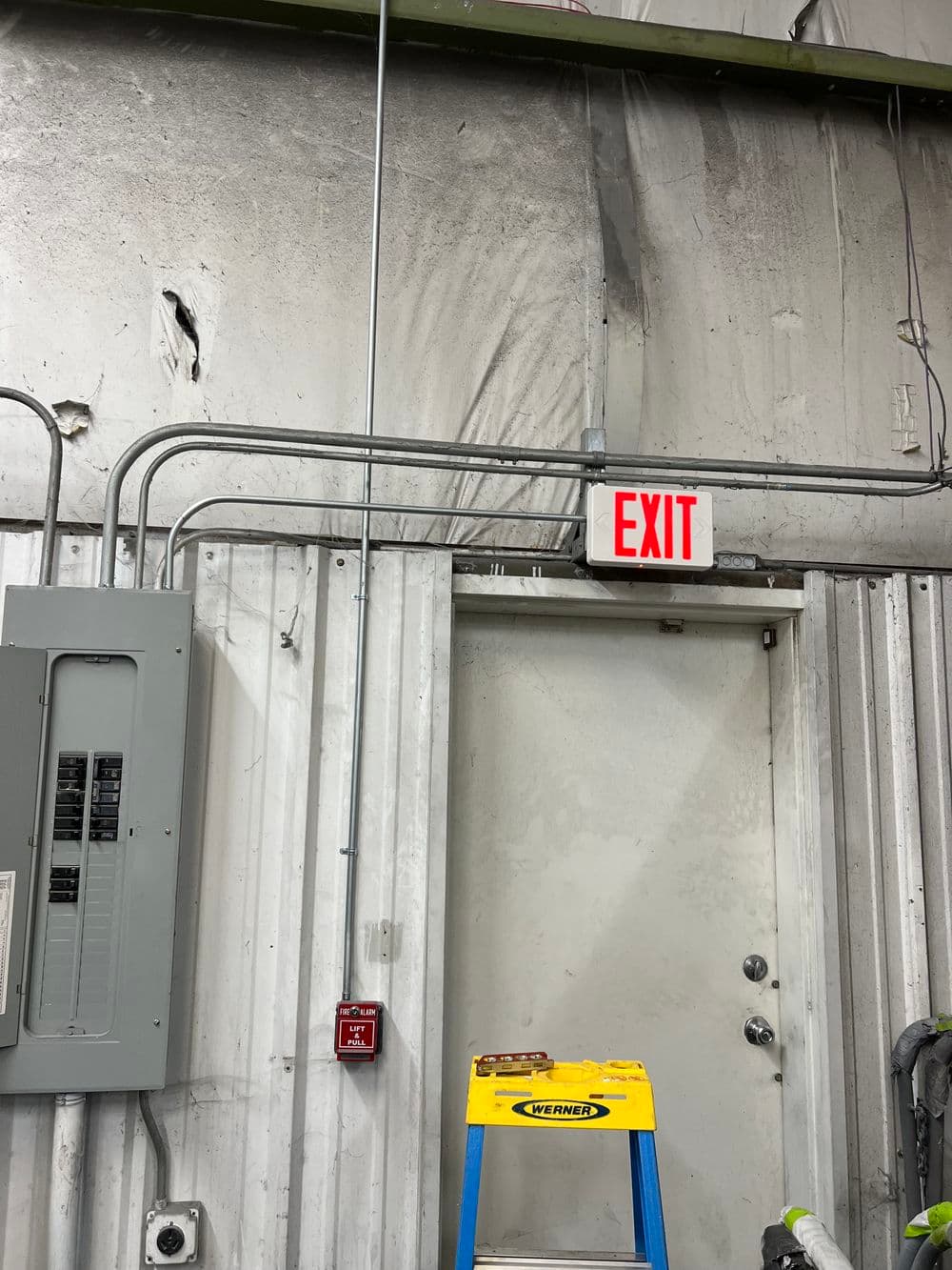 Warehouse exit sign above a door with electrical panel and ladder nearby.