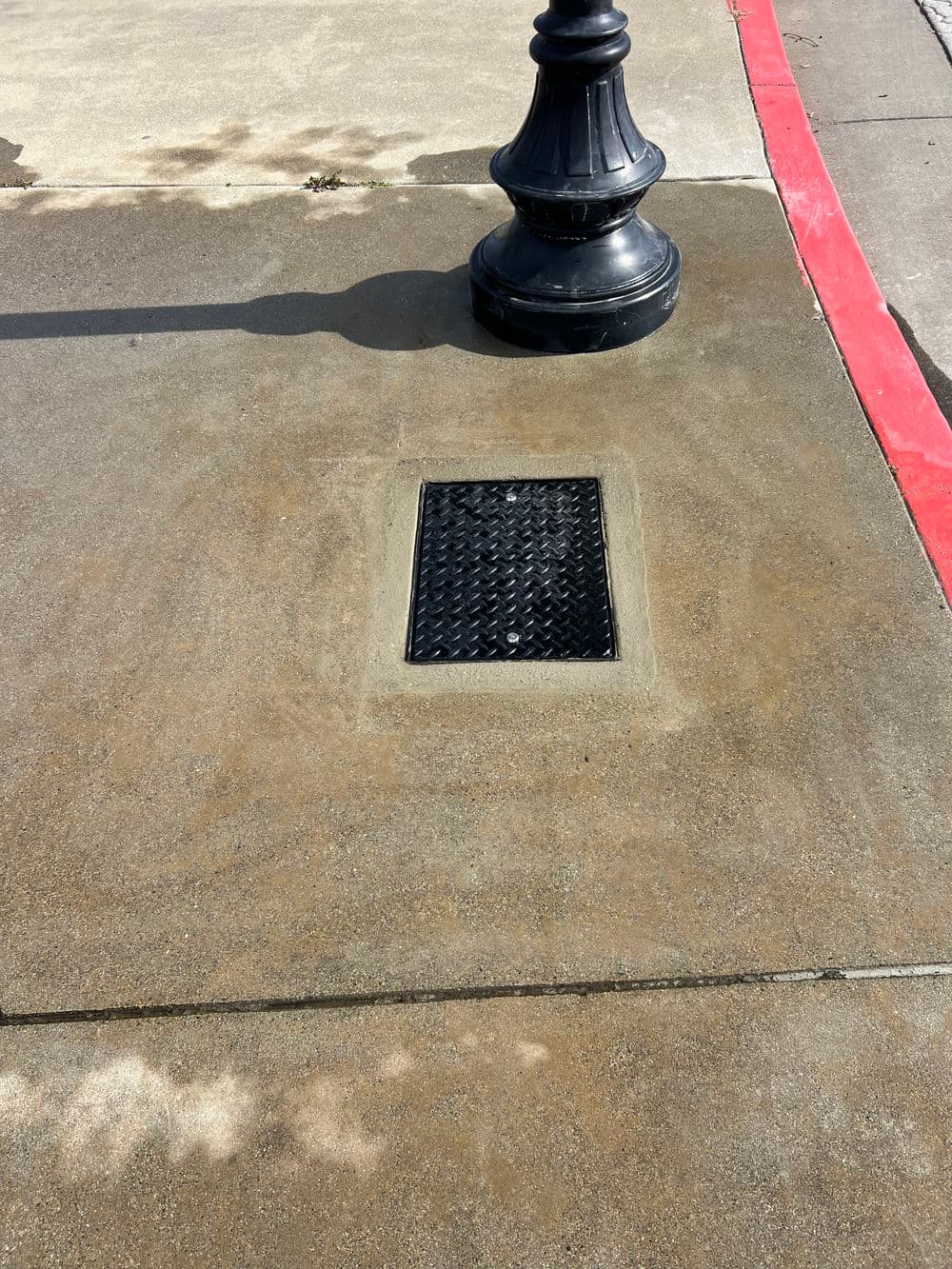 Black metal drain cover on a concrete sidewalk near a decorative street lamp.