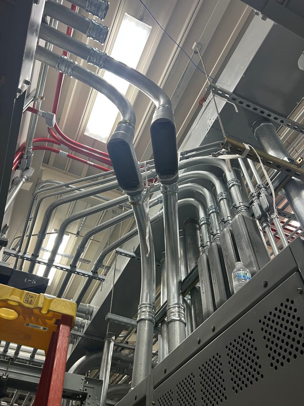 Industrial plumbing and ductwork with metal pipes and red wiring in a mechanical room.