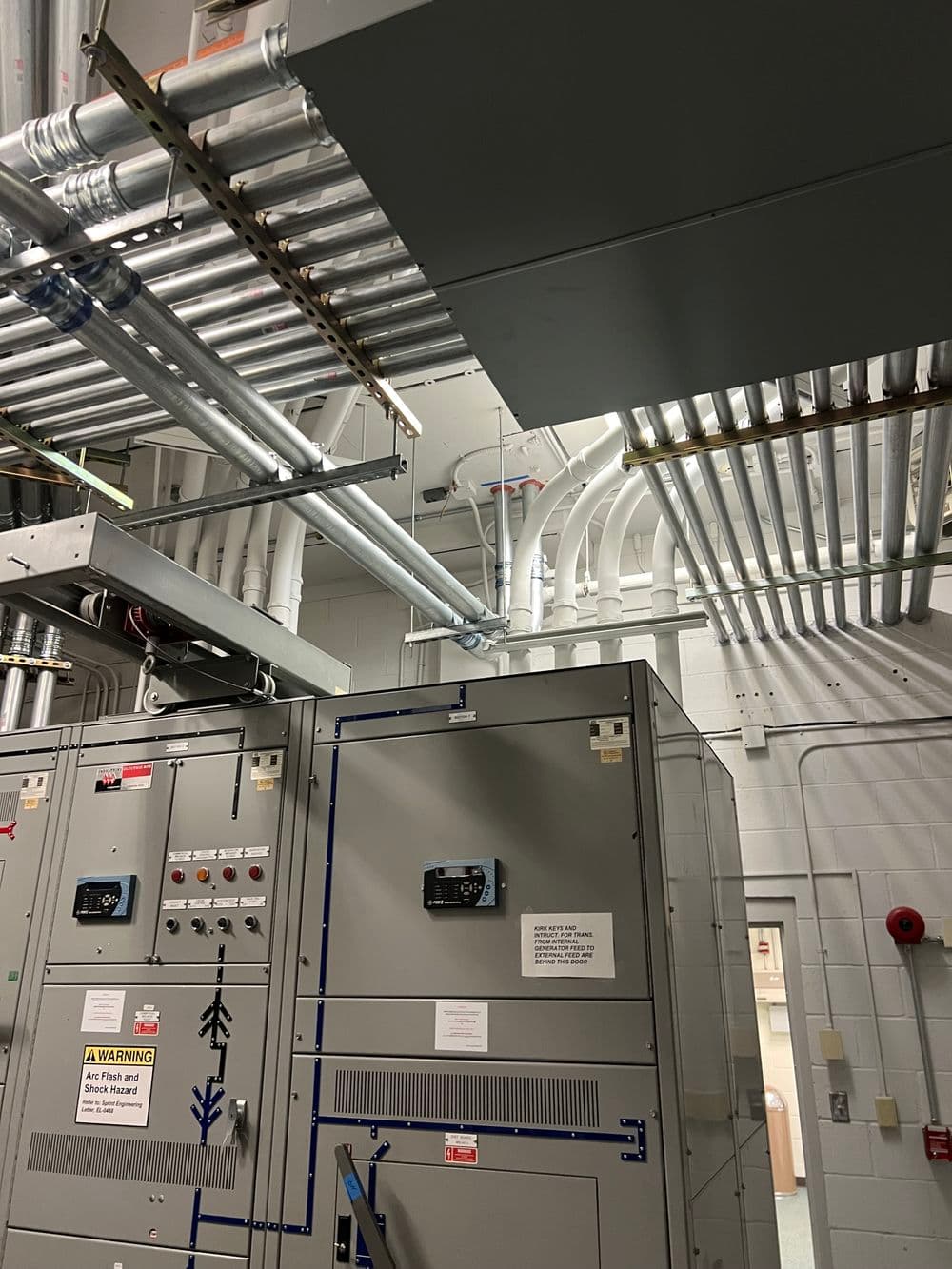 Industrial electrical equipment with ducts and pipes in a facility. Safety warning visible.