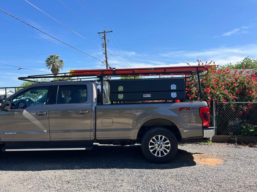 Silver pickup truck with a tool rack and storage box parked beside flowering bushes.